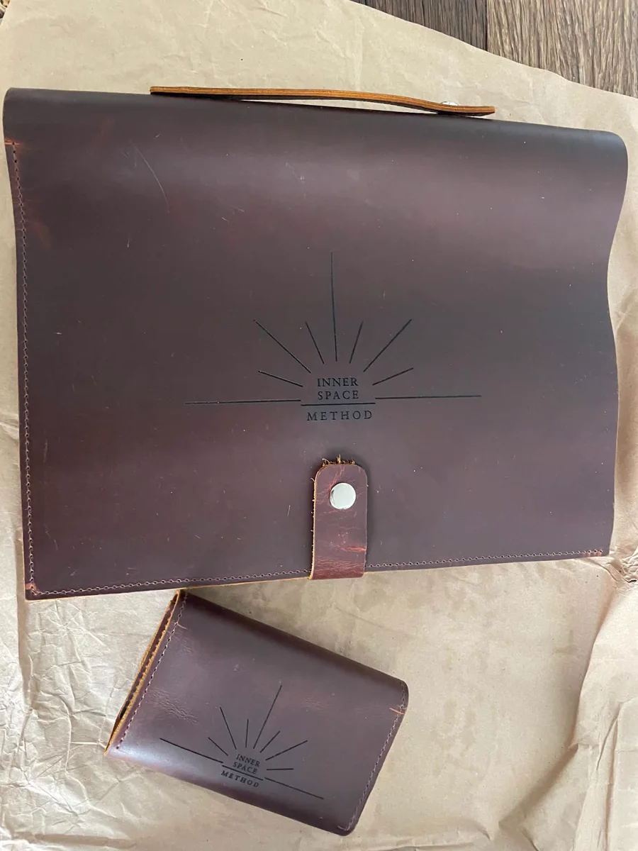 Das fertige Zeichen, in Leder geprägt: das strahlende Inner-Space-Method-Logo auf der Arbeitsmappe, daneben ein kleines Kartenetui. The finished mark, embossed in leather: the radiating Inner Space Method logo on the practice folio, a smaller card wallet beside it.