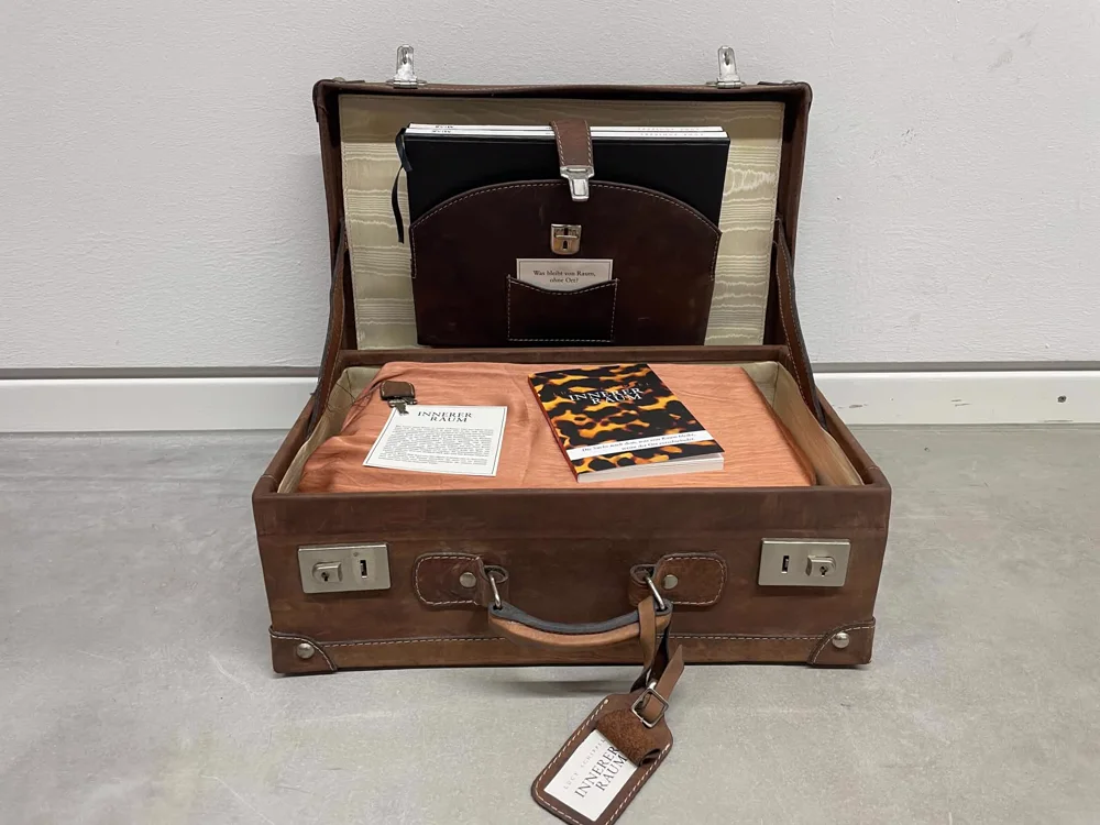 Die reisende Installation: ein offener Lederkoffer mit dem Buch INNERER RAUM, den fünf Phasen-Workbooks und einer Kraftpapier-Karte mit der Frage, was von Raum bleibt, wenn der Ort verschwindet. The travelling installation: an open leather suitcase holding the book INNERER RAUM, the five phase workbooks and a kraft card asking what remains of space without place.